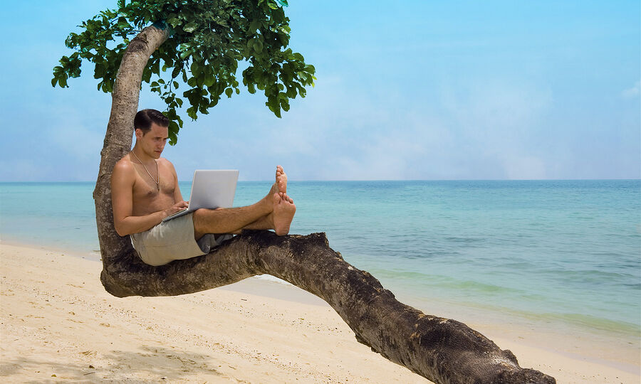 Attractive man with laptop seated in a tree on a tropical beach.